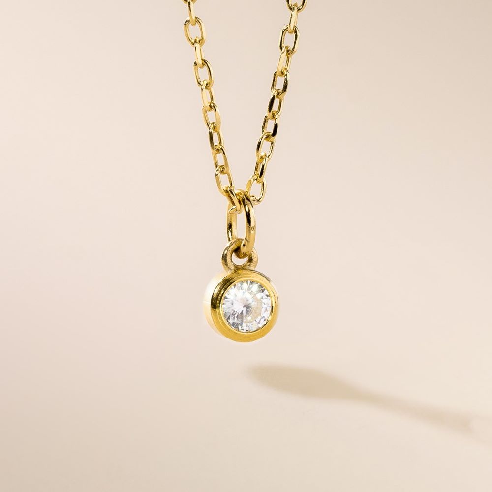 A gold pendant necklace with a single clear birthstone pendant, displayed against a beige background.