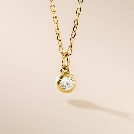 A gold pendant necklace with a single clear birthstone pendant, displayed against a beige background.