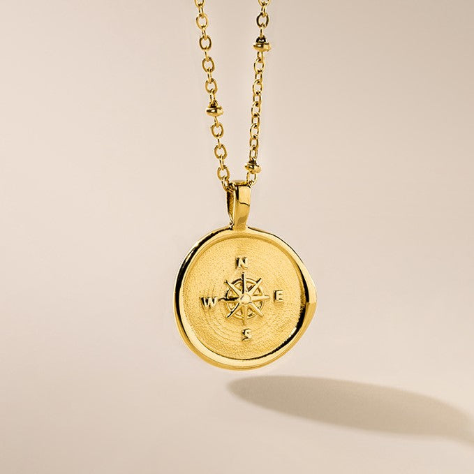 A gold-plated necklace with a round pendant featuring a compass design, displayed against a light background.