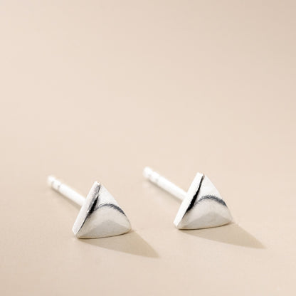 A pair of silver-colored fine triangle stud earrings on a beige background.