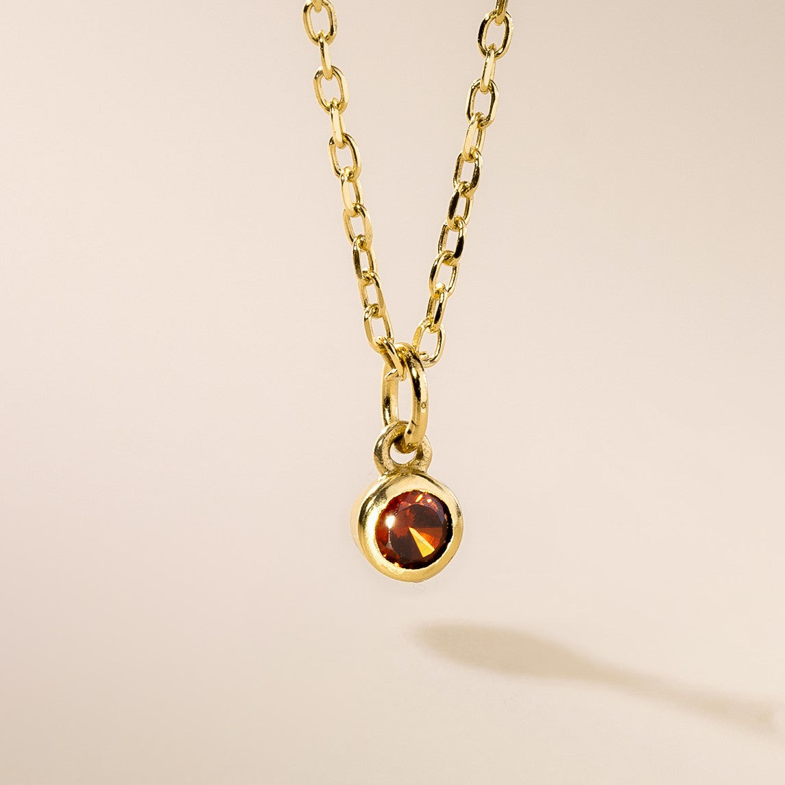 A gold pendant necklace with a round birthstone charm hanging from it, displayed against a neutral background.