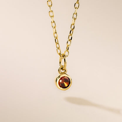 A gold pendant necklace with a round birthstone charm hanging from it, displayed against a neutral background.