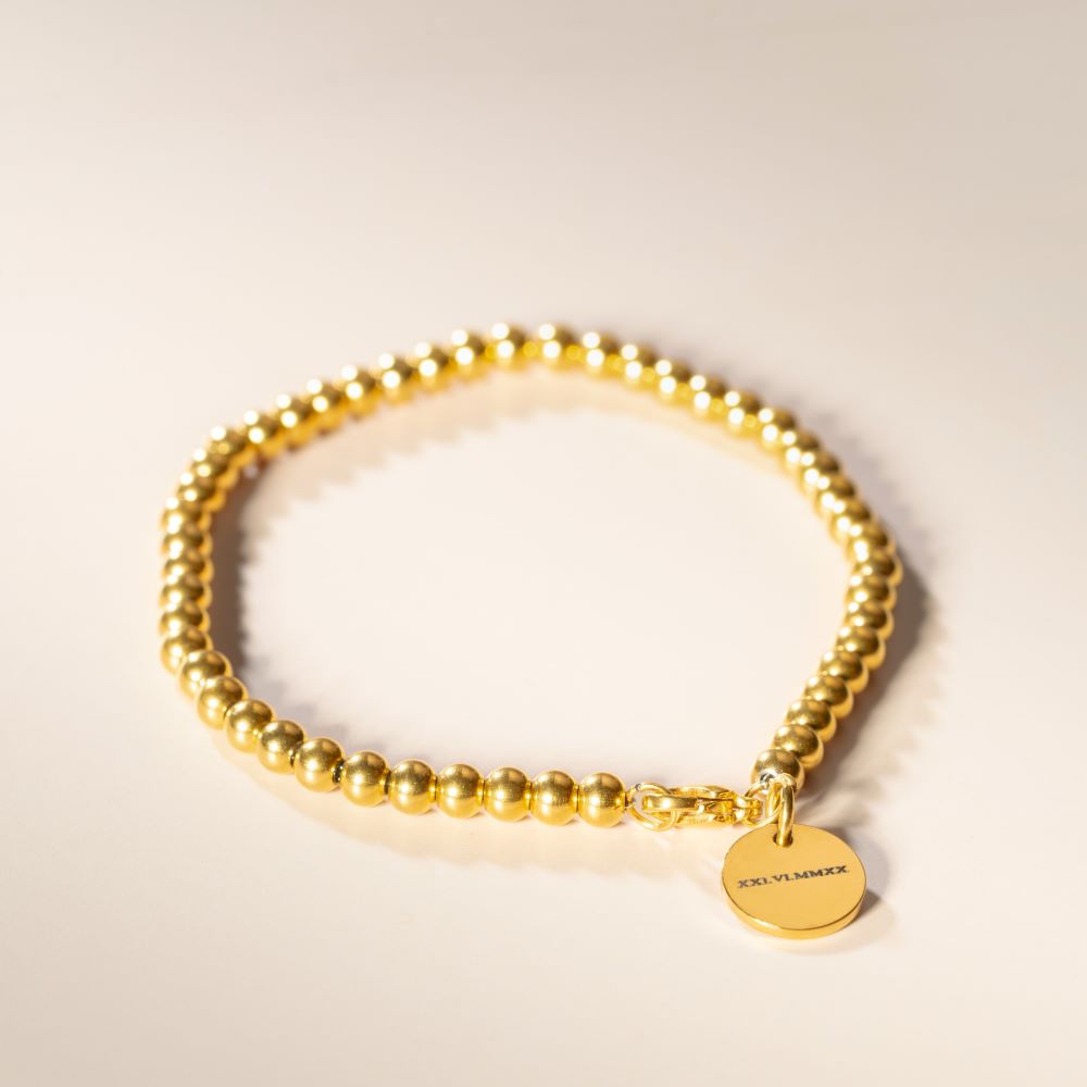 A gold-plated bracelet with a solid pattern and a circular pendant engraved with an initial, displayed against a neutral background.