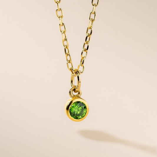 A gold pendant necklace with a round green birthstone charm hanging from it, displayed against a light background.