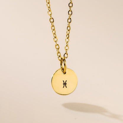 A gold-plated pendant necklace with the Pisces astrological sign engraved on a disc, attached to a gold-plated chain, presented against a neutral background.