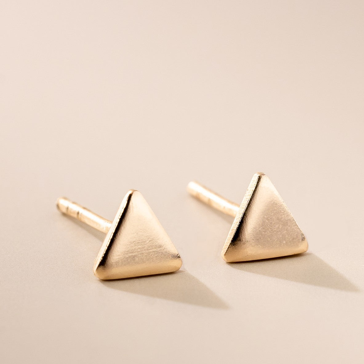 A pair of rose gold triangle stud earrings placed on a beige surface.