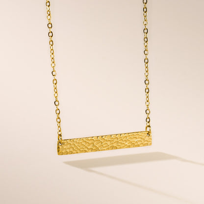 A gold hammered bar necklace with a chain and a clasp, displayed against a neutral background.