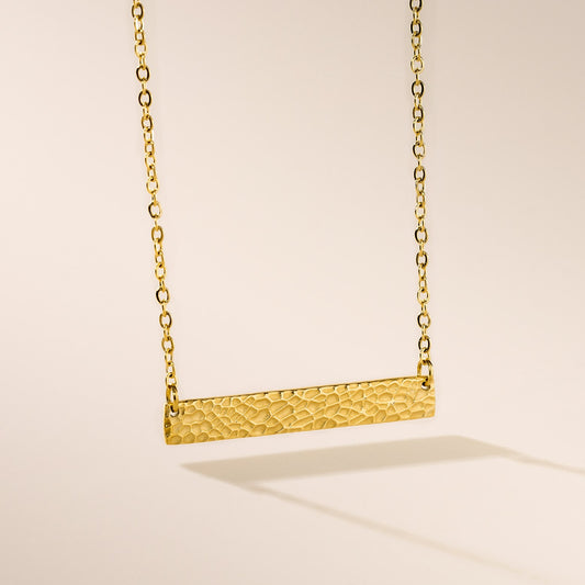 A gold hammered bar necklace with a chain and a clasp, displayed against a neutral background.