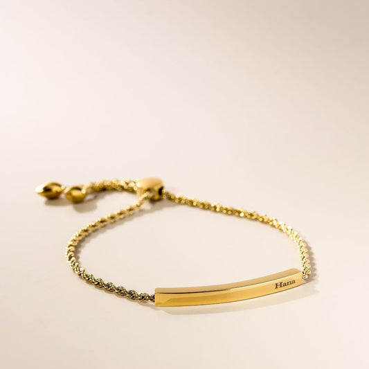 A gold-plated rope bracelet with an adjustable slider, featuring a bar for engraving, displayed against a neutral background.