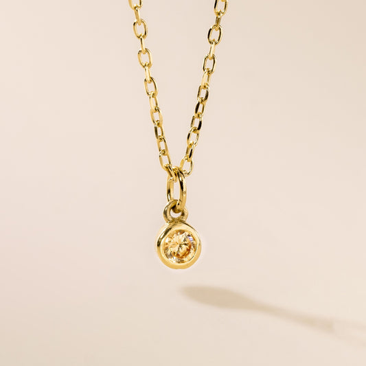 A gold pendant necklace with a single birthstone pendant hanging from a chain, presented against a light background.